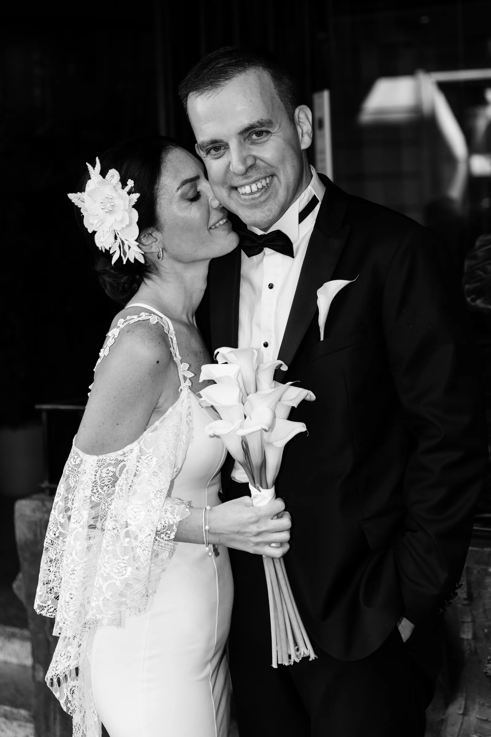 Black and white: the bride kisses the groom's cheek holding calla lilies outside the consulate