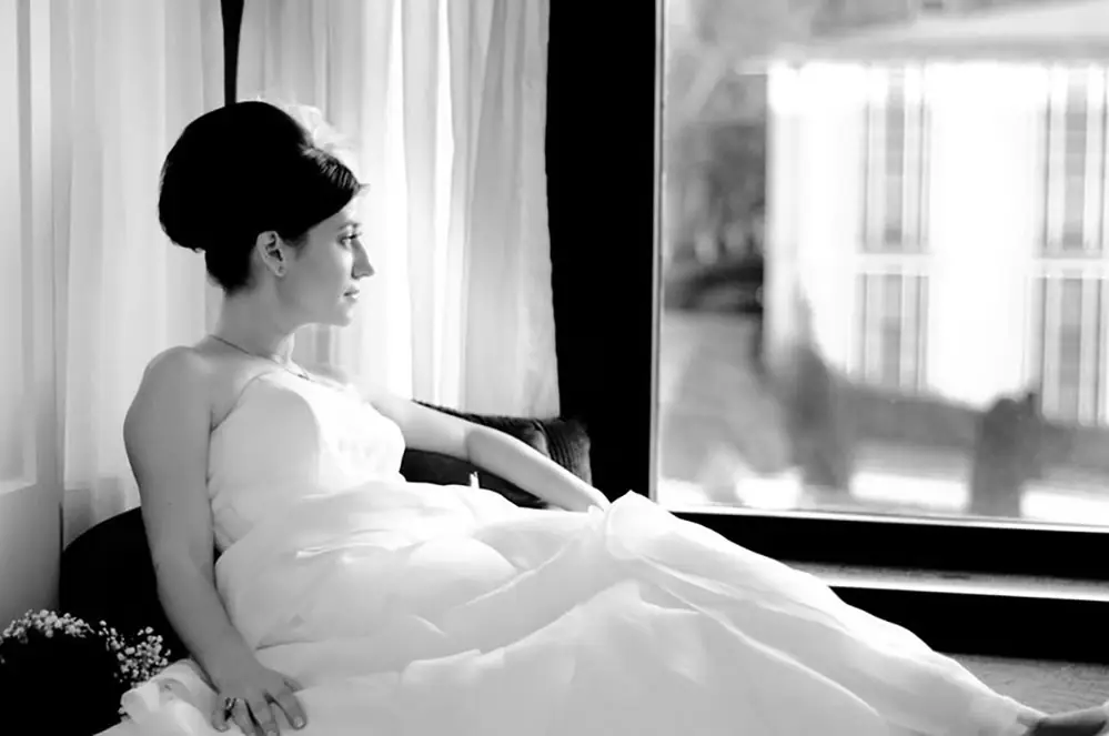 Bride in her gown waiting by a window in soft daylight, black and white