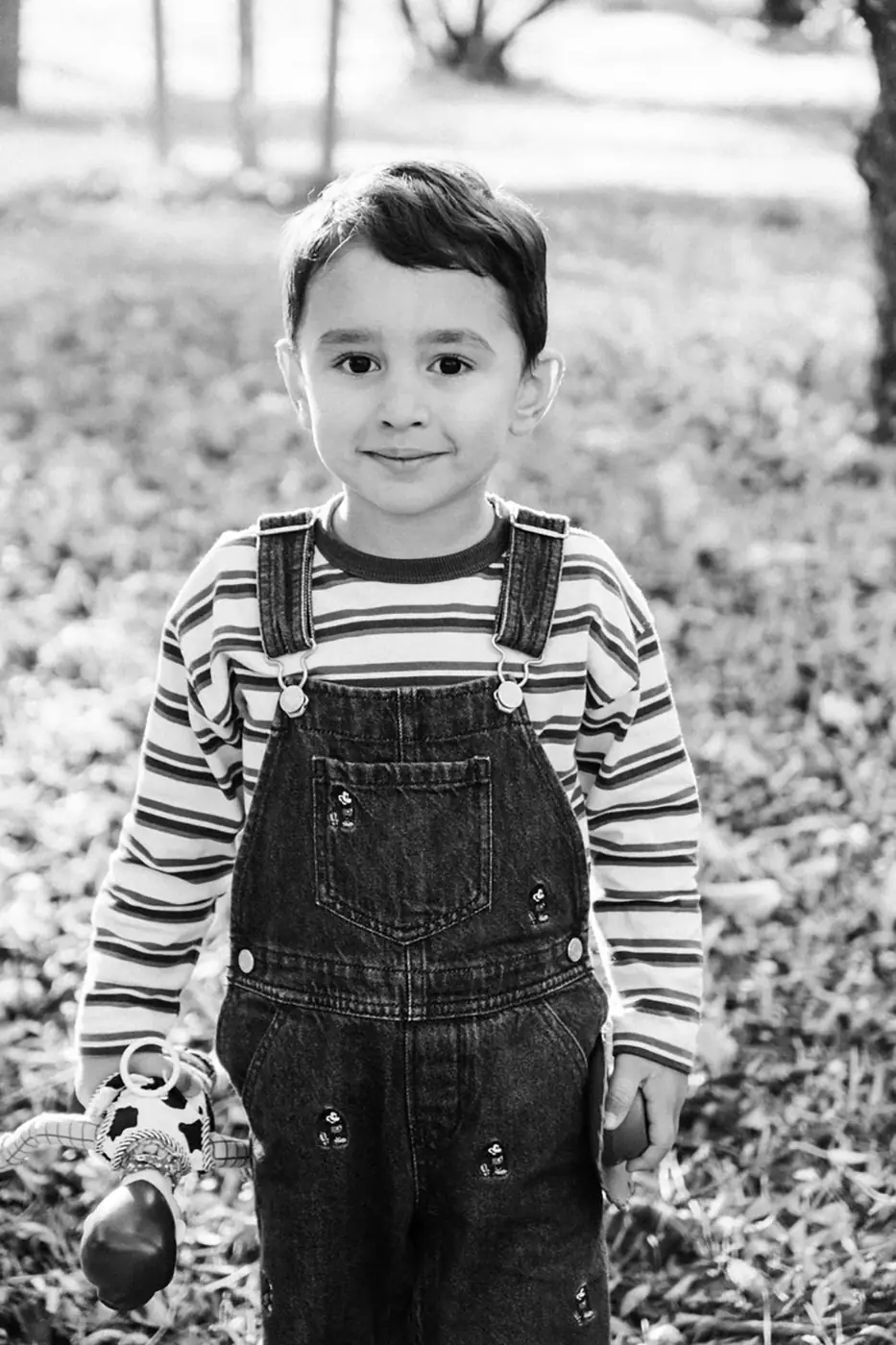 Boy in striped shirt and dungarees standing on a park path, black and white