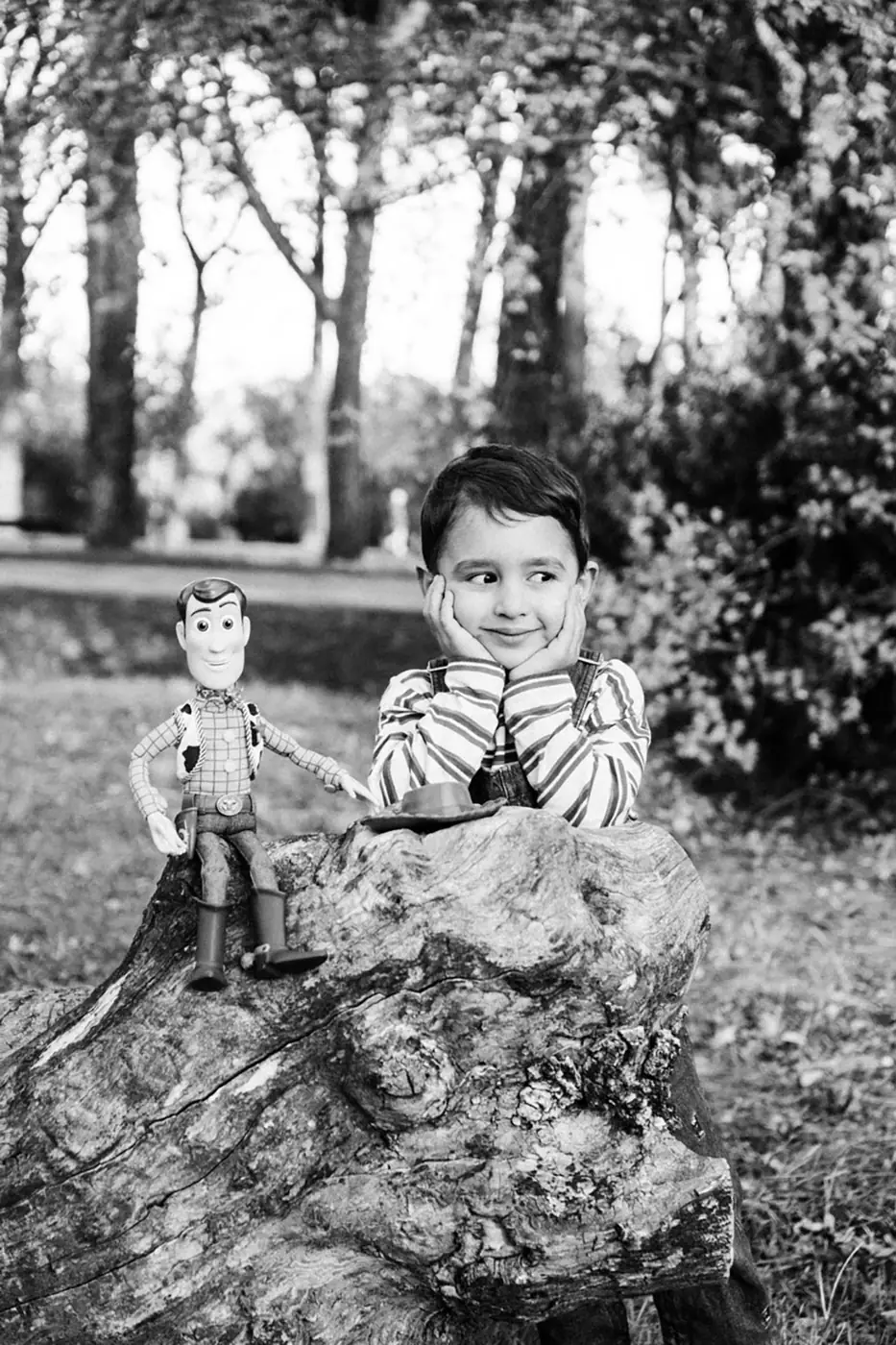 Boy resting his chin on his hands beside a toy cowboy on a tree stump, black and white