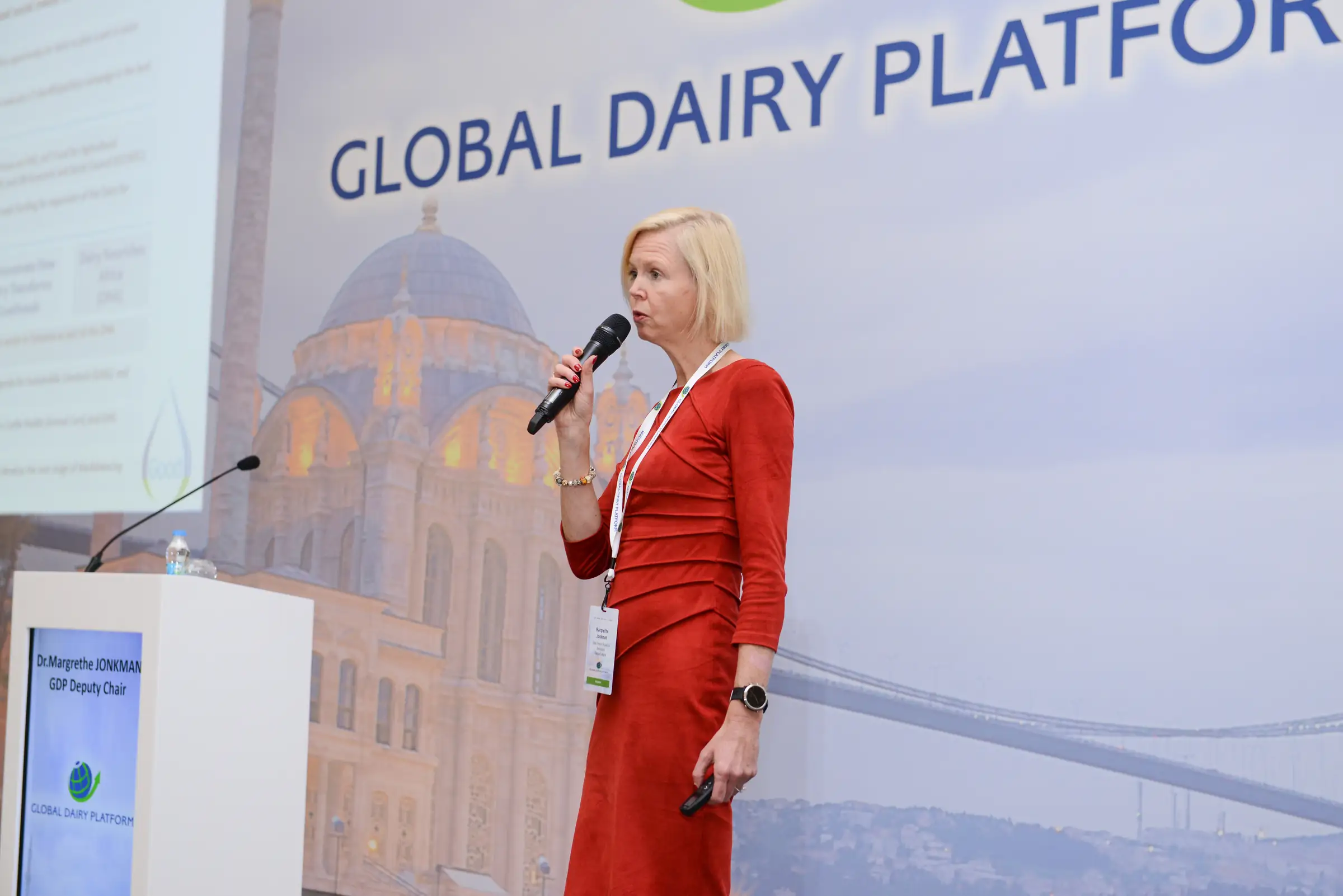 Speaker in a red suit addressing delegates with a handheld microphone at a corporate conference
