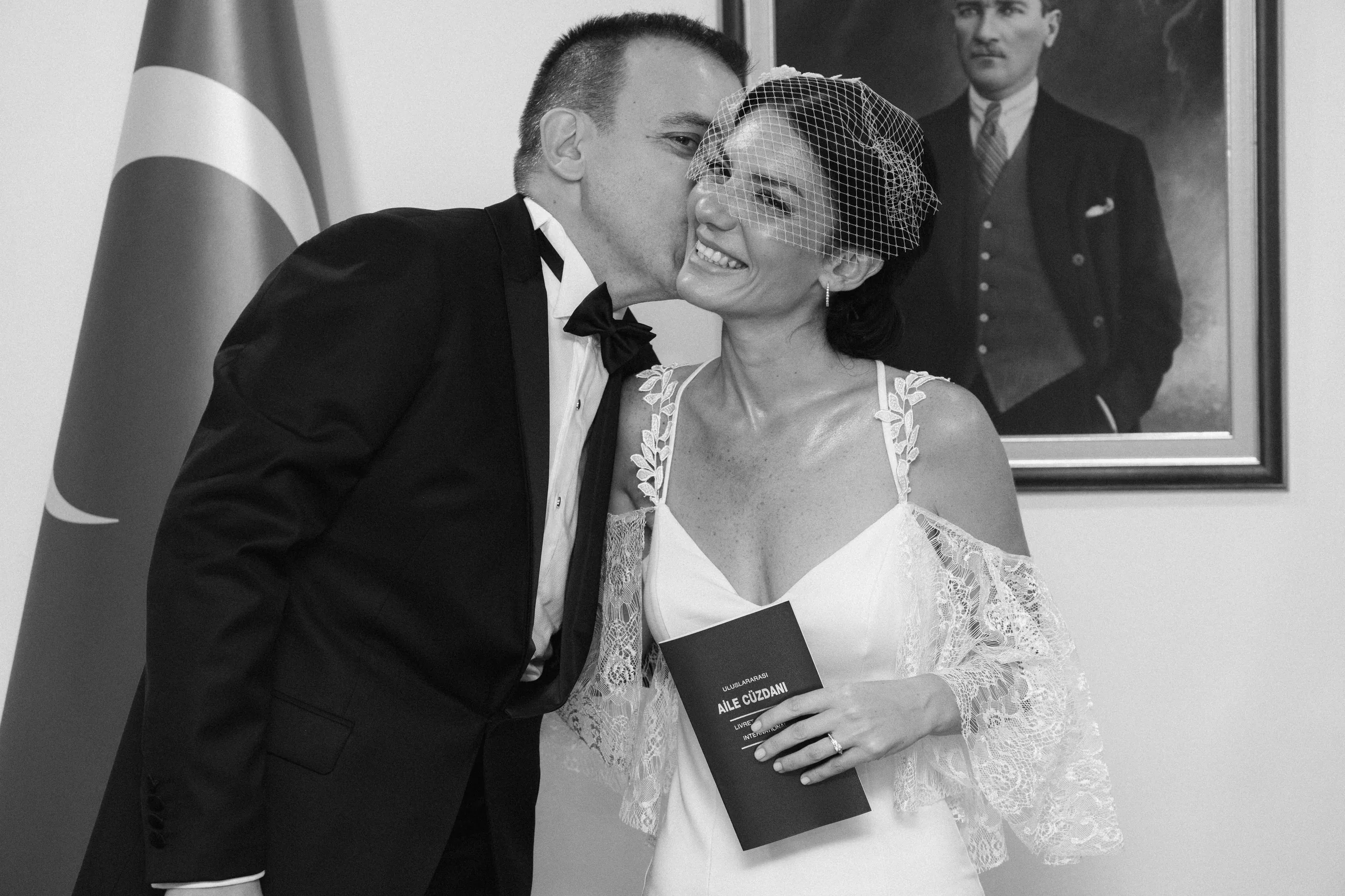Black and white: the groom kisses the bride on the cheek while she holds the family booklet