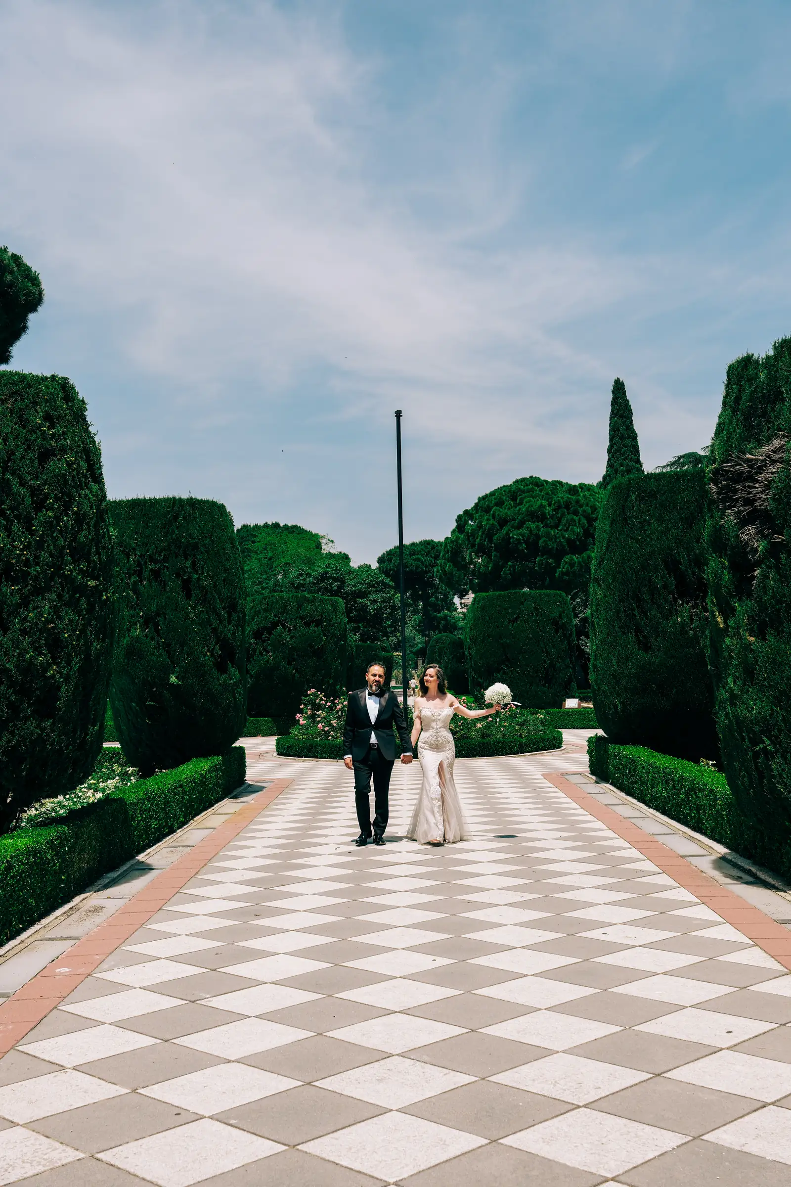 Bride and groom on the chequered stone path between clipped cypresses in Madrid