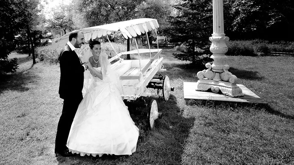 Bride and groom beside a vintage open car in a garden, black and white