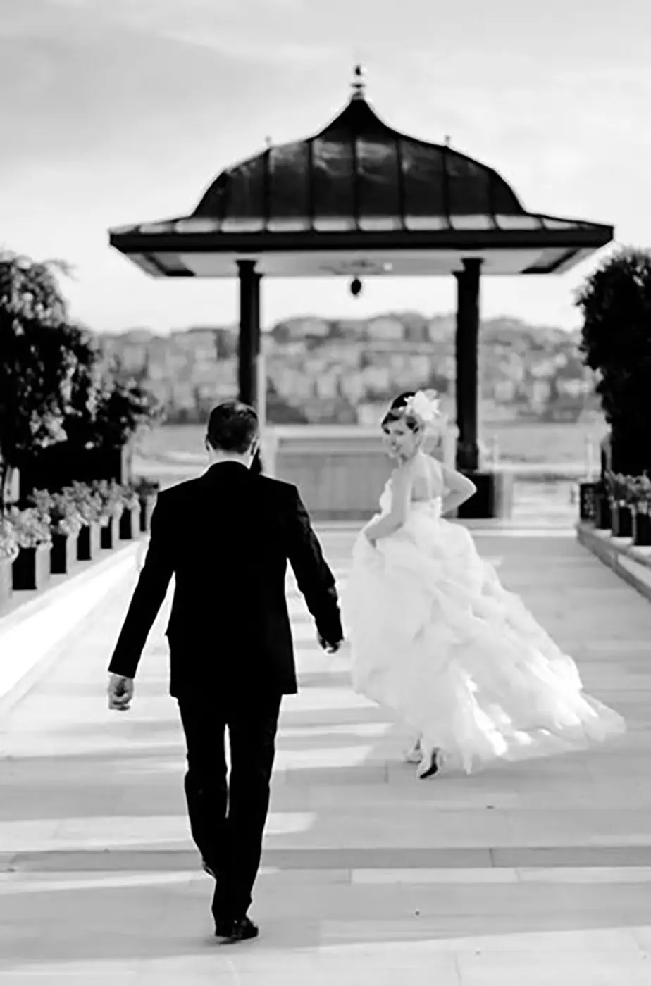 Bride and groom walking away towards a waterside pavilion, black and white