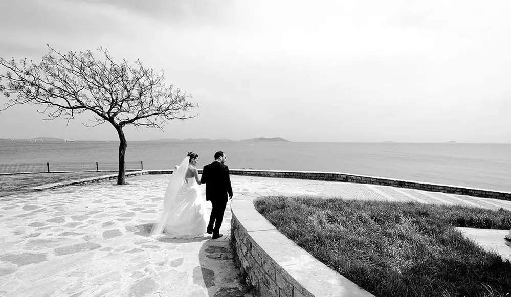 Couple walking a curved path beside a bare tree by the water, black and white
