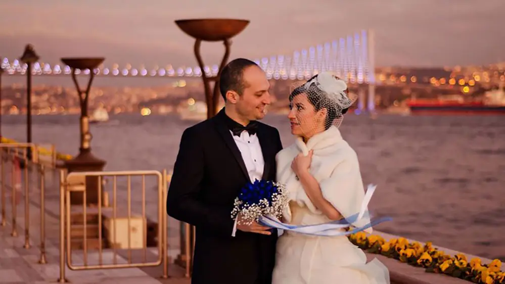 Bride and groom on a waterside terrace at night with a lit bridge behind them