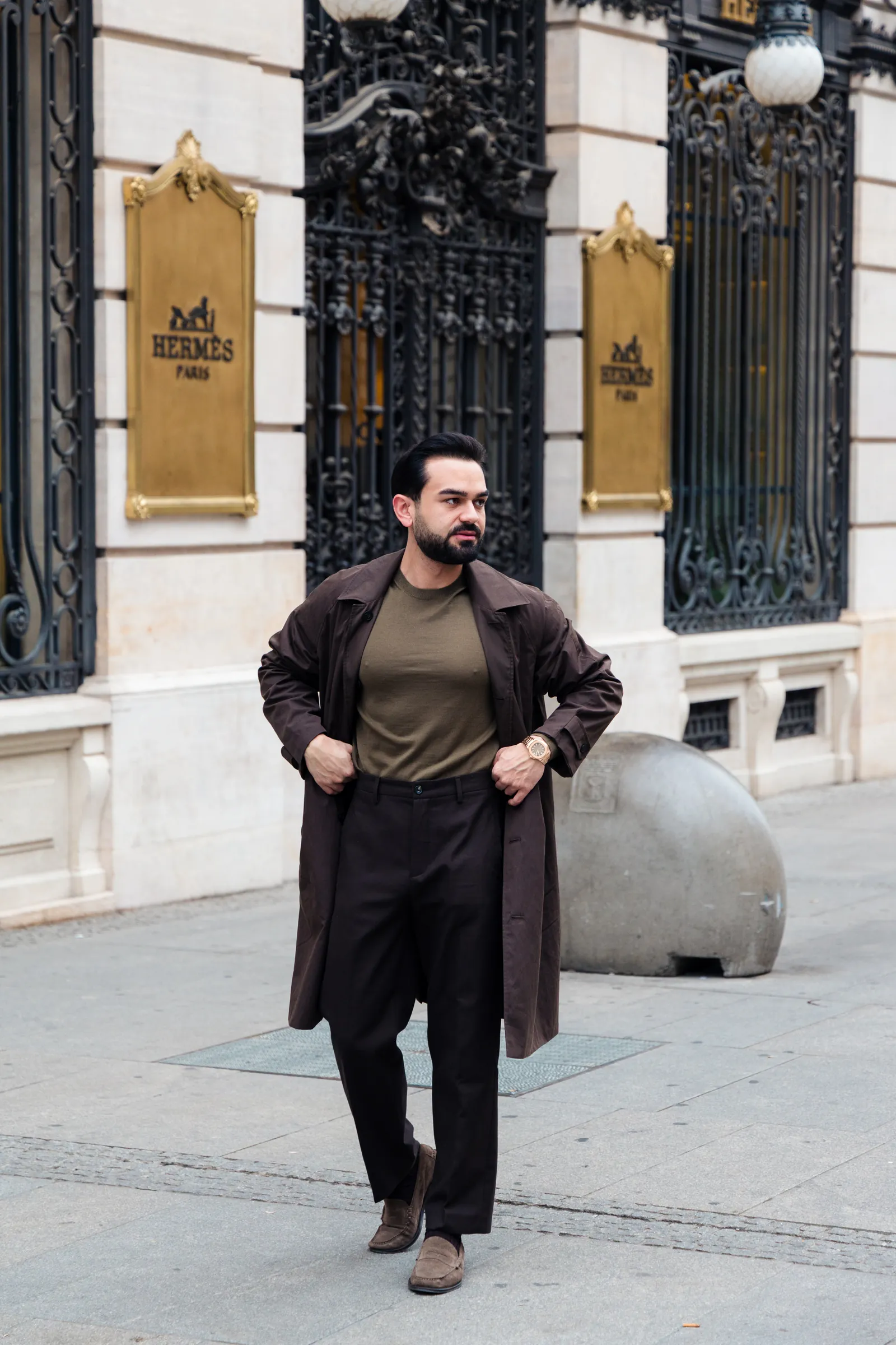 Man in a long brown coat over an olive knit in front of a stone facade with wrought iron windows
