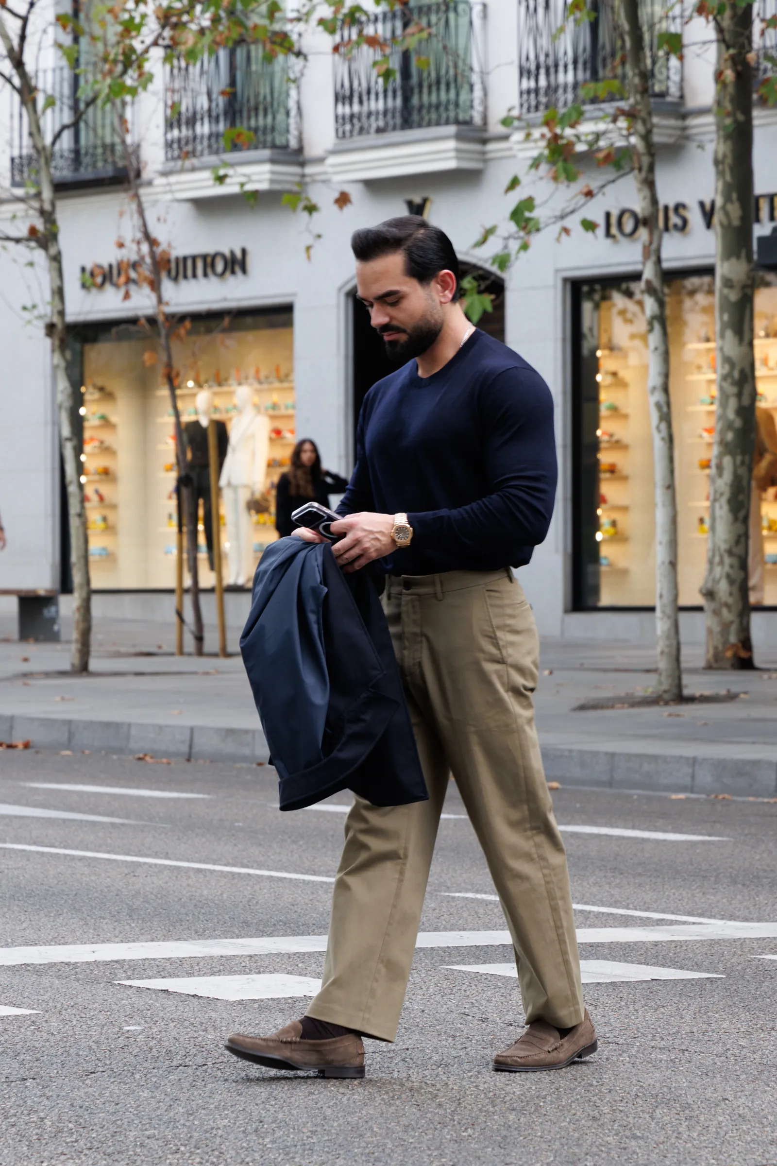 Man in a navy knit and sand trousers crossing a wide avenue in front of shop windows