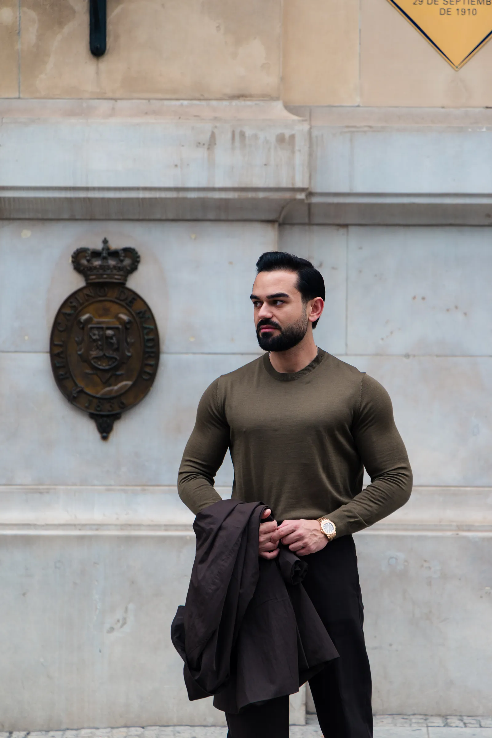 Man in an olive knit holding his coat beside a carved crest on a stone wall