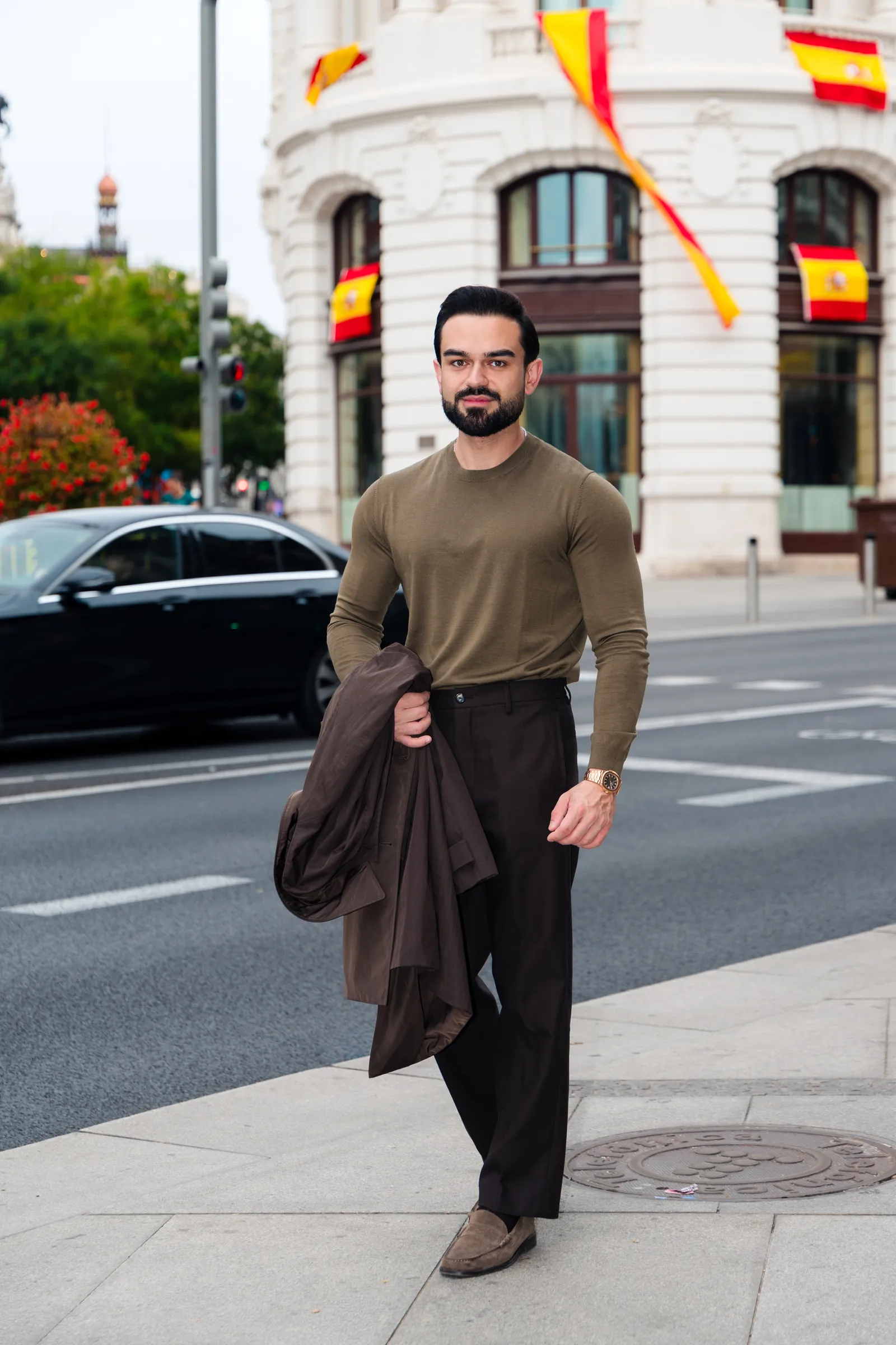 Man in an olive knit walking along a city street with Spanish flags on the building behind