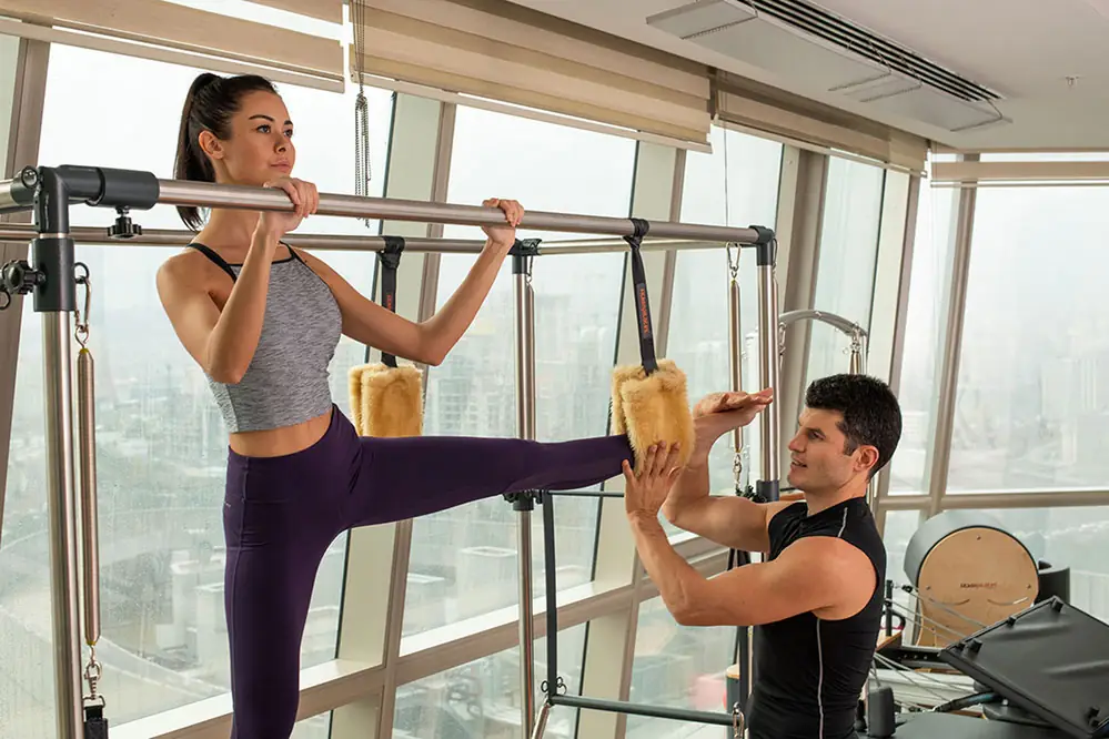 Trainer supporting a client through a standing pilates exercise beside a city window
